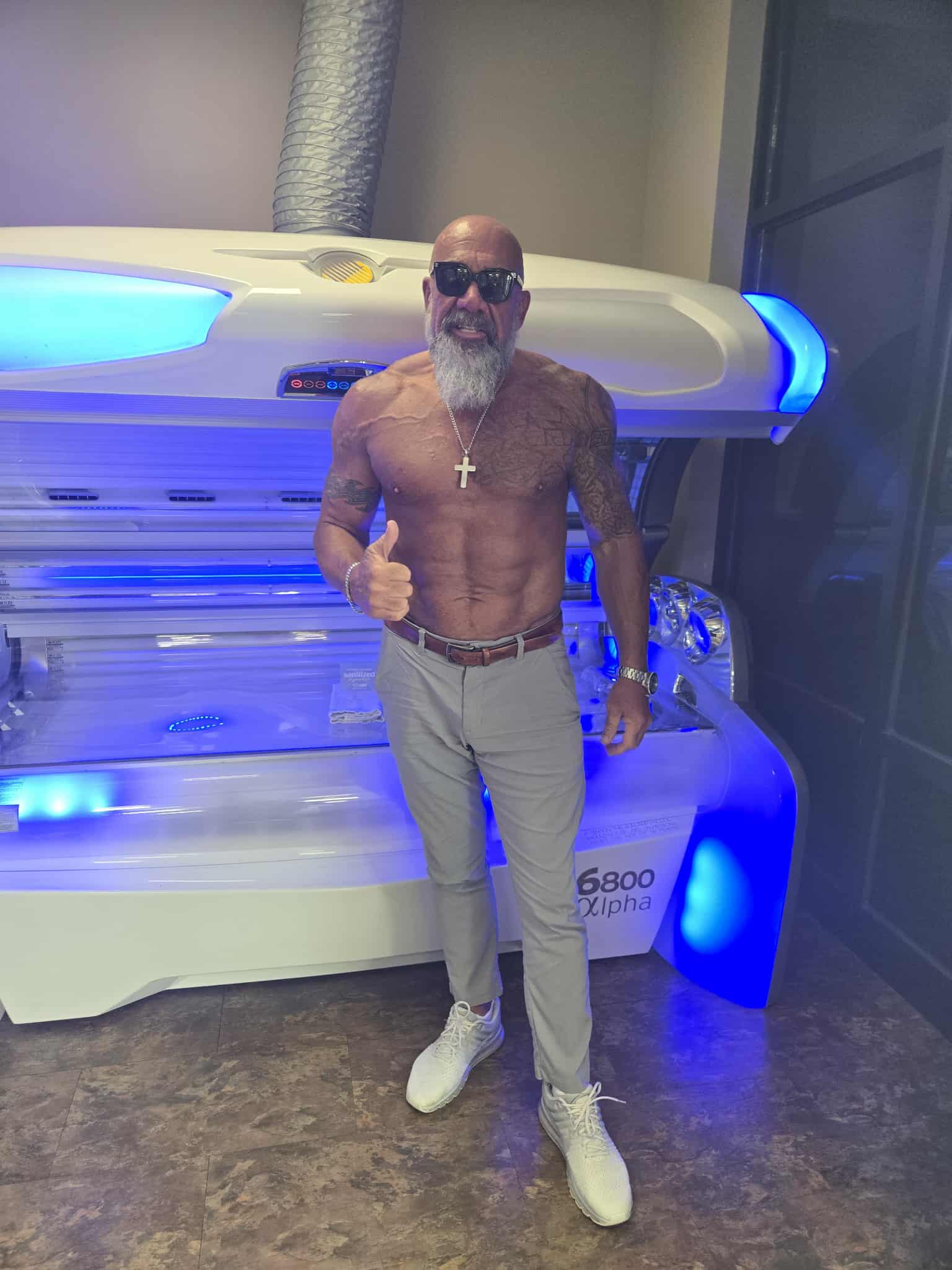 Hammer in beige pants and sunglasses giving a thumbs up next to tanning equipment with blue LED lighting